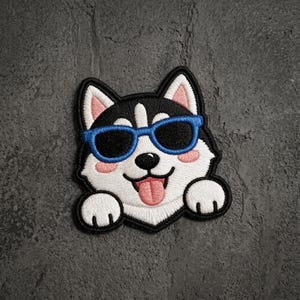 May include: An embroidered patch featuring a cartoon husky dog with black and white fur, wearing bright blue sunglasses. The dog has pink inner ears and cheeks, a black nose, and a pink tongue sticking out, with two white paws visible at the bottom.