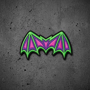 May include: An embroidered patch shaped like a pair of bat wings. The design features a vibrant neon green border and outlines, with bright pink sections filling the wing panels. The entire patch is finished with a thick black stitched edge.
