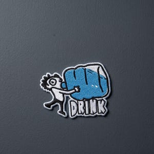 May include: An embroidered patch featuring a cartoon figure with black hair and white clothing holding a large blue jug to their mouth. Below the figure, the word DRINK is stitched in white block letters with a thick black outline on a white background.