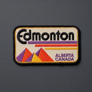 May include: A rectangular embroidered patch with a cream background and a black border. It features the word Edmonton in black text, with purple, orange, and yellow horizontal lines above stylized mountain peaks in the same colors. Alberta Canada is written in purple text.
