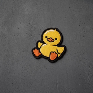 May include: An embroidered patch featuring a yellow duckling with orange feet and a beak, outlined in black thread. The duckling has large black eyes and a small white highlight on its head, creating a cartoon-style appearance with stitched details.