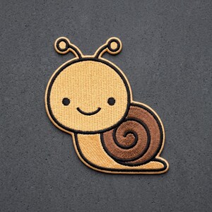 May include: An embroidered iron-on patch shaped like a snail with a light yellow body and a dark brown spiral shell. The snail features a simple black outline, two antennae with circular tips, two small black eyes, and a curved black mouth line.