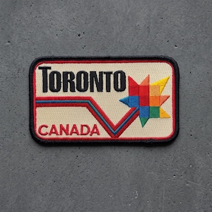 May include: A rectangular embroidered patch with a beige background and a black border. The text TORONTO is in black, and CANADA is in red. A multi-colored geometric maple leaf design in red, orange, yellow, blue, and green sits next to a red and blue line.