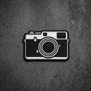 May include: A black and white embroidered patch shaped like a vintage camera. The design features a central circular lens, a rectangular viewfinder, and a top-mounted shutter button, all outlined with a thick black border and white stitching.