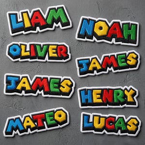 May include: A collection of eight embroidered fabric patches featuring the names LIAM, NOAH, OLIVER, JAMES, JAMES, HENRY, MATEO, and LUCAS. Each letter is rendered in a bold, blocky font with a multi-colored design of blue, yellow, red, and green.