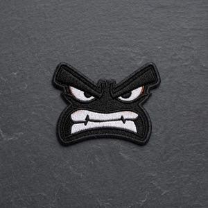 May include: An embroidered patch featuring a stylized, angry face with a black background, white eyes with orange accents, and a wide, white-toothed mouth. The design has a thick black border and a textured, stitched surface throughout the shape.