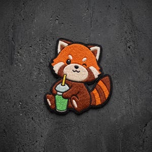 May include: An embroidered patch featuring a cartoon red panda with orange and brown fur, white facial markings, and a striped tail. The red panda is holding a green cup with a yellow straw, winking with one eye, and outlined in black thread.