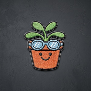 May include: An embroidered patch featuring a terracotta-colored flower pot with a smiling face, wearing light blue goggles. A green plant with four leaves sprouts from the top of the pot. The entire design is outlined in thick black thread.