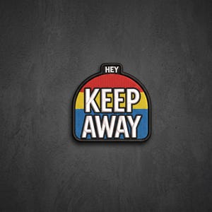 May include: A semi-circular embroidered patch with a black border featuring the text HEY KEEP AWAY in bold white letters. The background consists of three horizontal stripes in red, yellow, and blue, with a black tab at the top center.