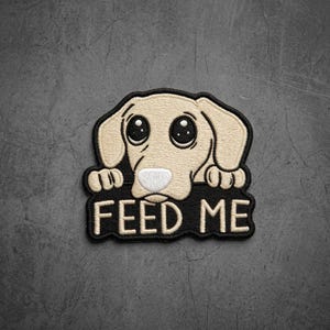 May include: An embroidered patch featuring a light beige puppy with large dark eyes and floppy ears resting its paws on a black base. The text FEED ME is stitched in bold beige capital letters across the bottom of the black section.