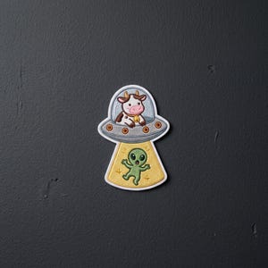 May include: An embroidered patch featuring a brown and white cow with a yellow bell sitting inside a grey flying saucer. A yellow beam of light extends from the bottom of the saucer, illuminating a small green alien with large black eyes.