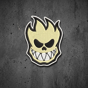May include: An embroidered patch featuring a stylized cream-colored skull with a flame-like top, black oval eyes, and a wide grin with sharp white teeth. The design is outlined in thick black and white borders, creating a high-contrast graphic.