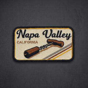May include: A rectangular embroidered patch with a cream-colored background and a black border. It features the black text Napa Valley and brown text CALIFORNIA. An embroidered brown corkscrew is depicted diagonally across the center.