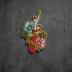 May include: An embroidered patch featuring a red seam ripper tool centered between a teal flower and a pink flower. A gold banner wraps around the tool with the black text No Regrets. The design uses detailed stitching in red, teal, pink, and gold.