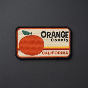 May include: A rectangular embroidered patch with a cream-colored background. It features a large orange fruit with a green leaf on the left. The text reads ORANGE County CALIFORNIA in black and orange, with horizontal yellow and orange stripes.