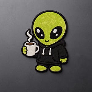 May include: An embroidered patch featuring a lime green alien character with large black eyes, wearing a black hooded sweatshirt with white drawstrings. The alien is holding a white mug filled with a dark brown beverage with white steam rising.