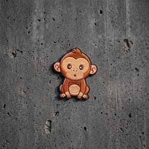 May include: An embroidered patch featuring a cartoon monkey with light brown fur on its face and belly, and darker brown fur on its head, ears, arms, and legs. The monkey has large dark eyes, a small nose, and a surprised expression with an open mouth.