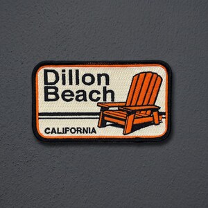 May include: A rectangular embroidered patch with a cream background and a black border. It features the black text Dillon Beach and CALIFORNIA, along with an illustration of an orange Adirondack chair set against a black horizontal line.