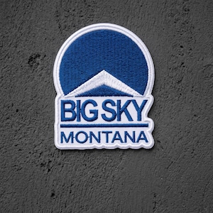 May include: An embroidered patch featuring a dark blue semi-circle top with a white mountain peak graphic. Below the mountain, the text BIG SKY is embroidered in blue, followed by a horizontal line and the word MONTANA in blue lettering.