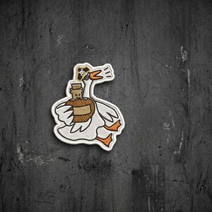 May include: An embroidered patch featuring a white goose with orange feet and beak, wearing a small black pirate hat with a white skull and crossbones emblem. The goose is holding a brown bottle with a cork stopper in its wing.