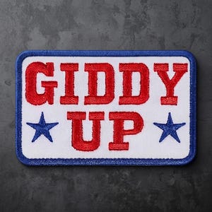 May include: A rectangular embroidered patch with a white background and a blue border. The text GIDDY UP is stitched in red capital letters. Two blue five-pointed stars are positioned on either side of the word UP, completing the design.