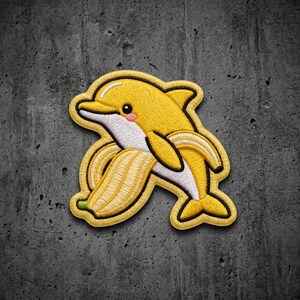 May include: An embroidered patch featuring a yellow dolphin with a white belly and a small pink cheek. The dolphin is partially emerging from a peeled yellow banana skin with a green stem tip, outlined in dark thread against a textured background.
