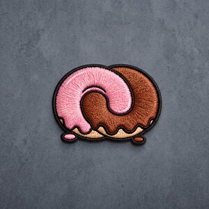 May include: An embroidered patch shaped like a pretzel, featuring one half covered in pink icing and the other half in brown chocolate icing, with a black stitched border outlining the entire shape and a light tan dough base visible at the bottom.