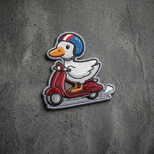 May include: An embroidered patch featuring a white duck wearing a blue, white, and red helmet while riding a red motor scooter. The duck has an orange beak and feet. A small white cloud of exhaust is visible behind the scooter on a gray background.