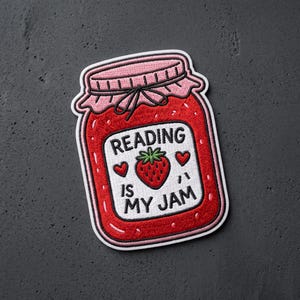 May include: A fabric patch shaped like a glass jar filled with red strawberry jam. The label features a red strawberry, two small red hearts, and the black embroidered text Reading Is My Jam. The jar has a pink lid with a black tie.