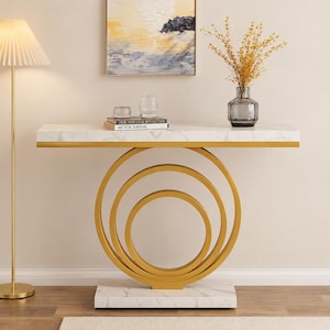 May include: A modern console table with a white marble top and gold-colored metal frame. The base features three concentric circles. The table is decorated with books, a vase with dried flowers, and a piece of abstract art.