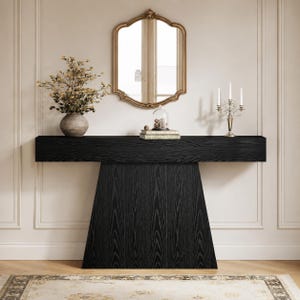 May include: A black wooden console table with a geometric base. A gold-framed mirror hangs above the table, which is decorated with a vase of flowers, books, and a decorative candle holder. The table has a modern design.