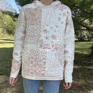 May include: A long-sleeve quilted hoodie featuring a patchwork design of cream and light pink fabric with various floral patterns. The garment includes a front kangaroo pocket and a hood, constructed with a stitched texture throughout the fabric.