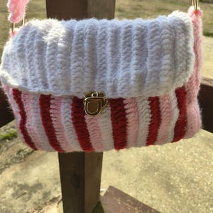 Pode incluir: Uma bolsa de crochê feita à mão com uma aba branca e um corpo com listras vermelhas e rosa. A bolsa tem um fecho dourado e alças rosa. A bolsa está suspensa em frente a uma grade de madeira.