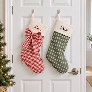 Puede incluir: Dos calcetines de Navidad colgados en una puerta blanca. El calcetín izquierdo presenta un patrón de cuadros vichy rojo y blanco con un lazo grande a juego y la palabra Mom bordada en rojo. El derecho es verde oliva con rayas verticales blancas y la palabra Dad bordada en negro.