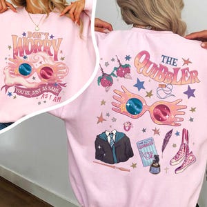 Può includere: Una felpa rosa chiaro con un design grafico. La parte anteriore presenta il testo "Don't Worry, You're Just As Sane As I Am" e un paio di occhiali. Il retro ha il testo "The Quibbler" e altre grafiche.