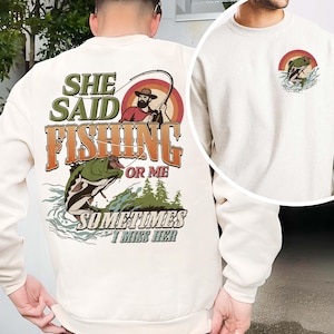 Könnte beinhalten: Cremefarbenes Sweatshirt mit einem Angel-Motiv. Auf der Rückseite befindet sich ein Design mit einem Fischer, einem Regenbogen und einem springenden Fisch, zusammen mit dem Text "She Said Fishing Or Me Sometimes I Miss Her". Eine kleinere Version des Designs befindet sich auf der Vorderseite.