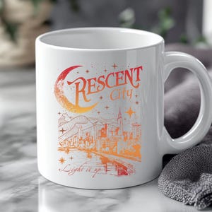 Puede incluir: Taza de cerámica blanca con una ilustración colorida. El diseño presenta el texto "Crescent City" sobre un paisaje urbano con una luna creciente y estrellas. Las palabras "Light it up" están debajo.