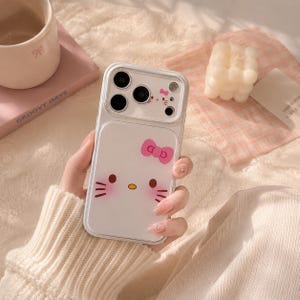 A person is holding a Hello Kitty phone case with a bow on it, surrounded by various items such as a mug, a candle, and a book.