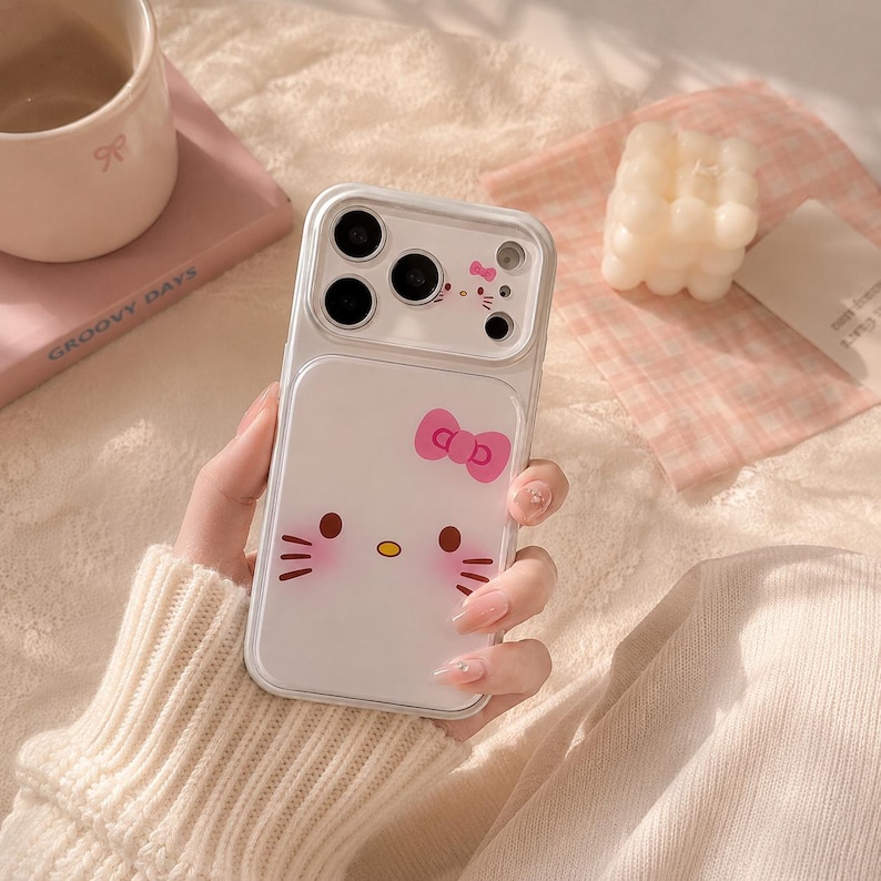 A person is holding a Hello Kitty phone case with a bow on it, surrounded by various items such as a mug, a candle, and a book.