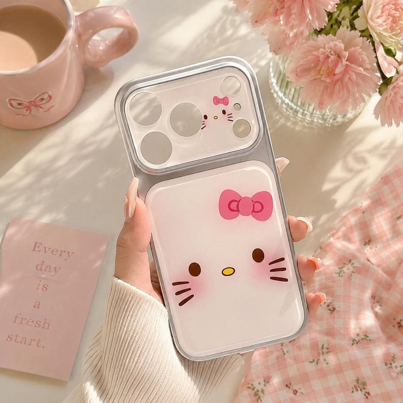 A hand holding a Hello Kitty phone case with a bow, surrounded by a pink mug, a vase of pink flowers, and a pink card with the phrase "Every day is a fresh start."