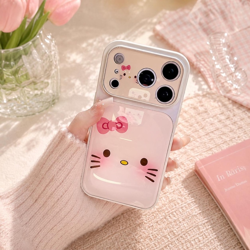 A person's hand holding a Hello Kitty phone case with a camera attached to it, along with a vase of pink tulips and a book.