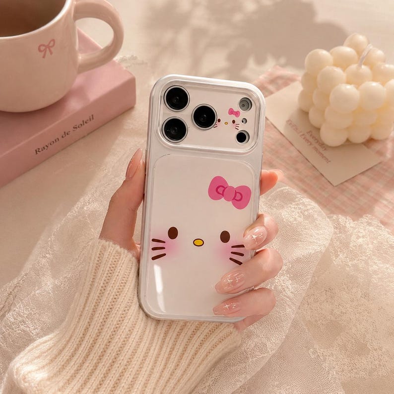 A person's hand holding a Hello Kitty phone case with a bow on it, surrounded by various items such as a mug, a candle, and a book.