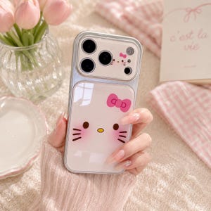 A person's hand holding a Hello Kitty phone case with a camera attached to it, along with a vase of pink flowers and a book in the background.