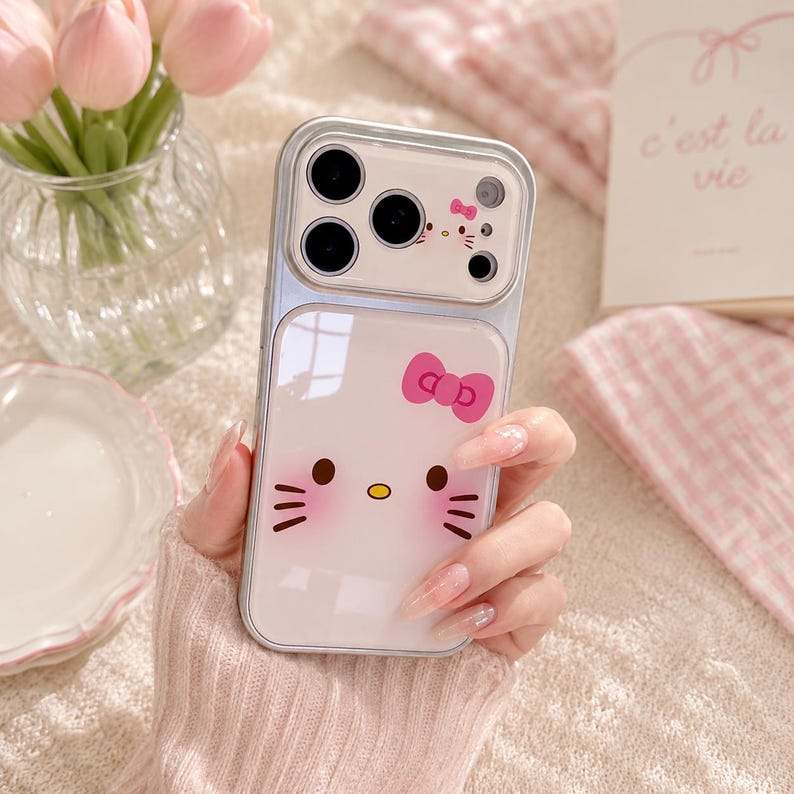 A person's hand holding a Hello Kitty phone case with a camera attached to it, along with a vase of pink flowers and a book in the background.