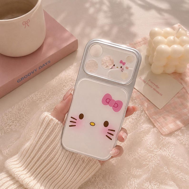 A hand holding a Hello Kitty phone case with a pink bow, surrounded by various items such as a mug, a book, and a candle.