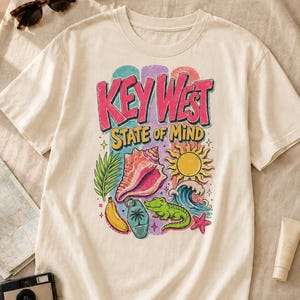 Peut inclure: T-shirt crème avec un graphisme coloré indiquant "KEY WEST STATE OF MIND". Le graphisme comprend un coquillage, un soleil, des vagues et un alligator vert. Des lunettes de soleil et un passeport sont visibles.