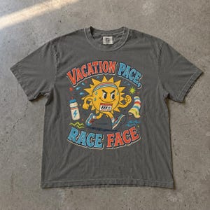 Könnte beinhalten: Ein anthrazitfarbenes Kurzarm-T-Shirt mit einer Grafik eines laufenden Sonnen-Charakters, der ein Stirnband und Turnschuhe trägt. Der Text lautet VACATION PACE, RACE FACE. Eine Wasserflasche und ein Handtuch sind neben der Sonne abgebildet.