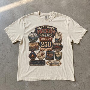 Puede incluir: Camiseta color crema con un diseño gráfico. El diseño presenta el texto "Patchwork Patriot Road Trip America 250" y parches temáticos de viajes. Los parches incluyen montañas, un coche y un motel.