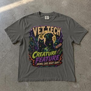 Pode incluir: Camiseta cinza com um gráfico colorido com um gato e um cão pretos, um estetoscópio e as palavras "VET TECH CREATURE FEATURE ANIMAL CARE NIGHT SHIFT". O design inclui raios e uma lua cheia.