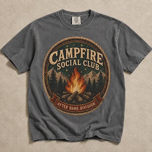 May include: A gray t-shirt with a circular graphic featuring a campfire scene. The graphic includes the text "CAMPFIRE SOCIAL CLUB" and "AFTER DARK DIVISION". The design incorporates a bonfire, trees, and a rope border.